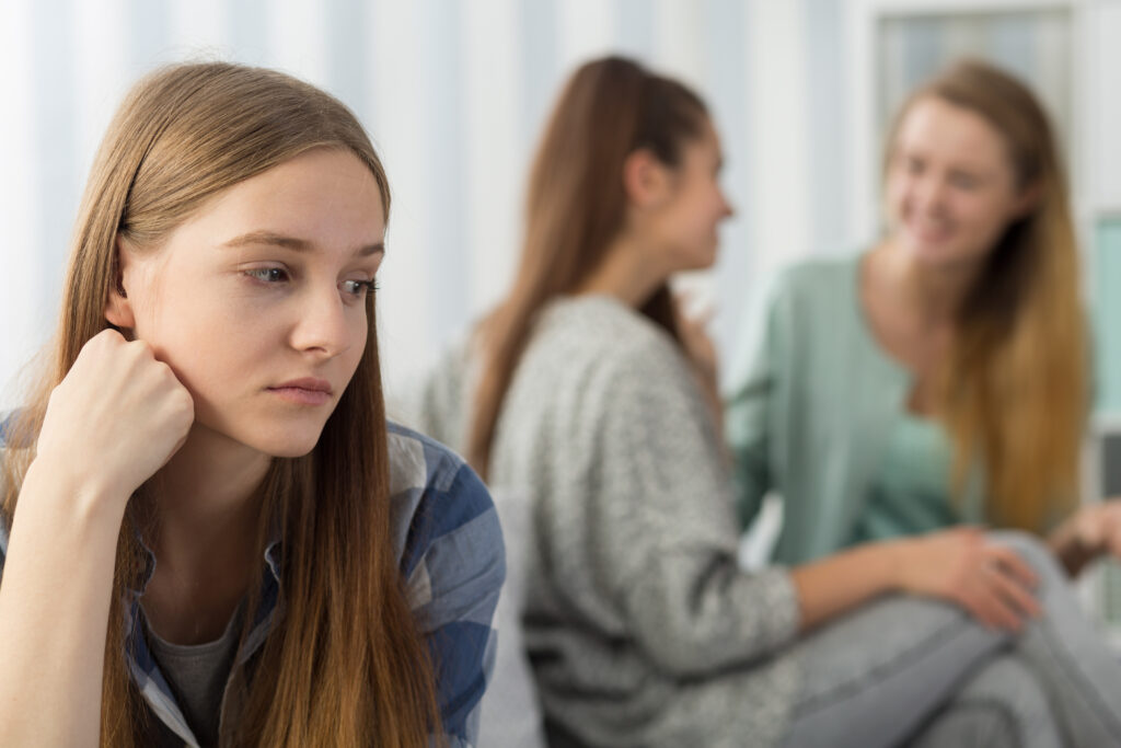 Das Bild zeigt 3 Frauen, wovon sich zwei, abgewandt, miteinander unterhalten. Die Frau im Vordergrund schaut nachdenklich.